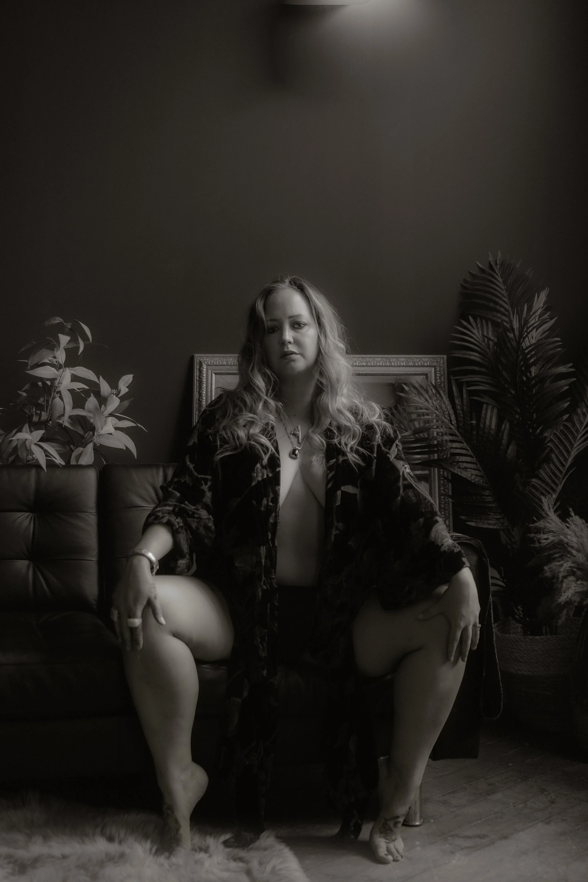 A black and white photo of a woman sitting on a couch with a serious expression, surrounded by plants in a dimly lit room.