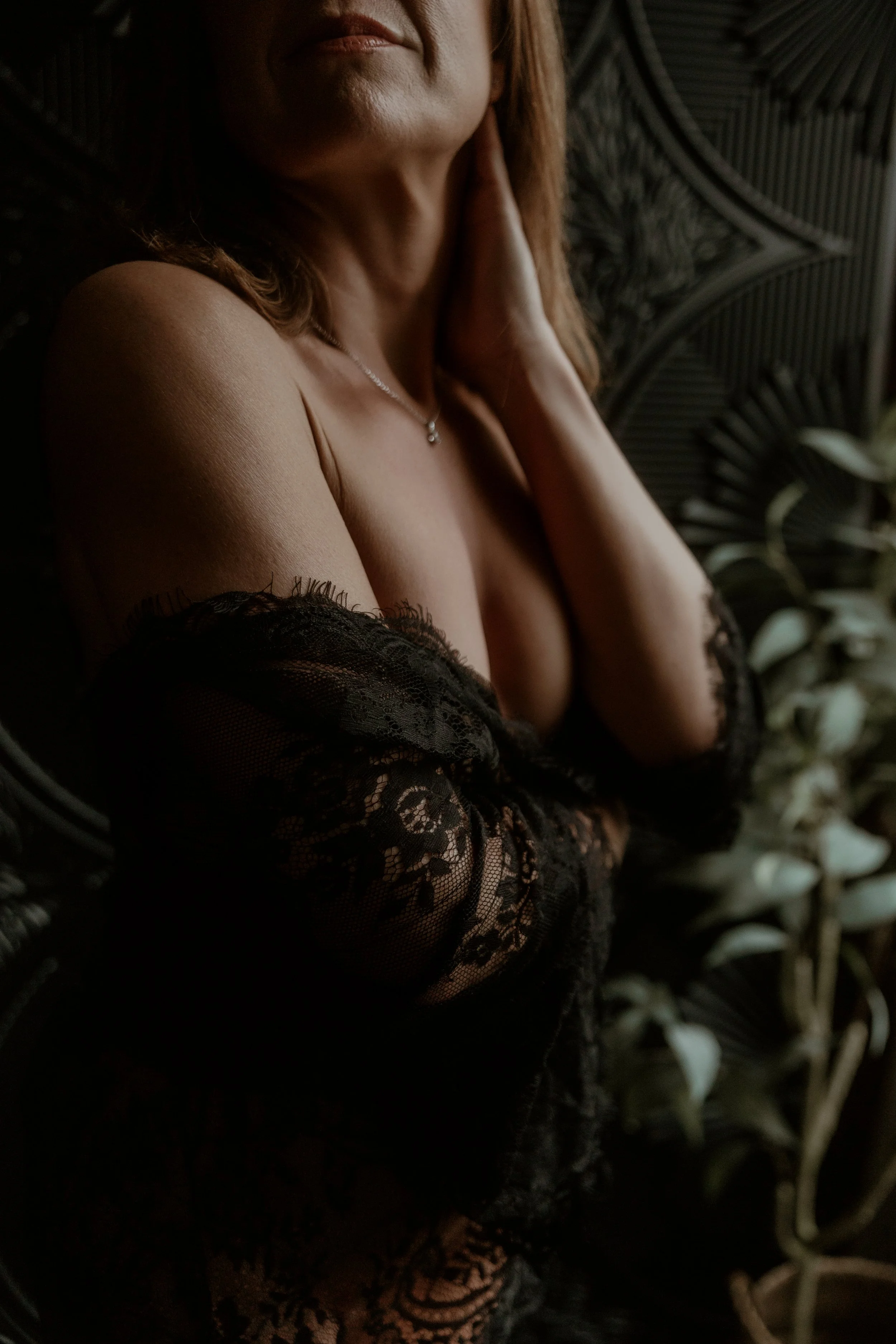 A close-up of a woman with her face partially visible, wearing a black lace cover-up, with a necklace, set against a dark background with plants.