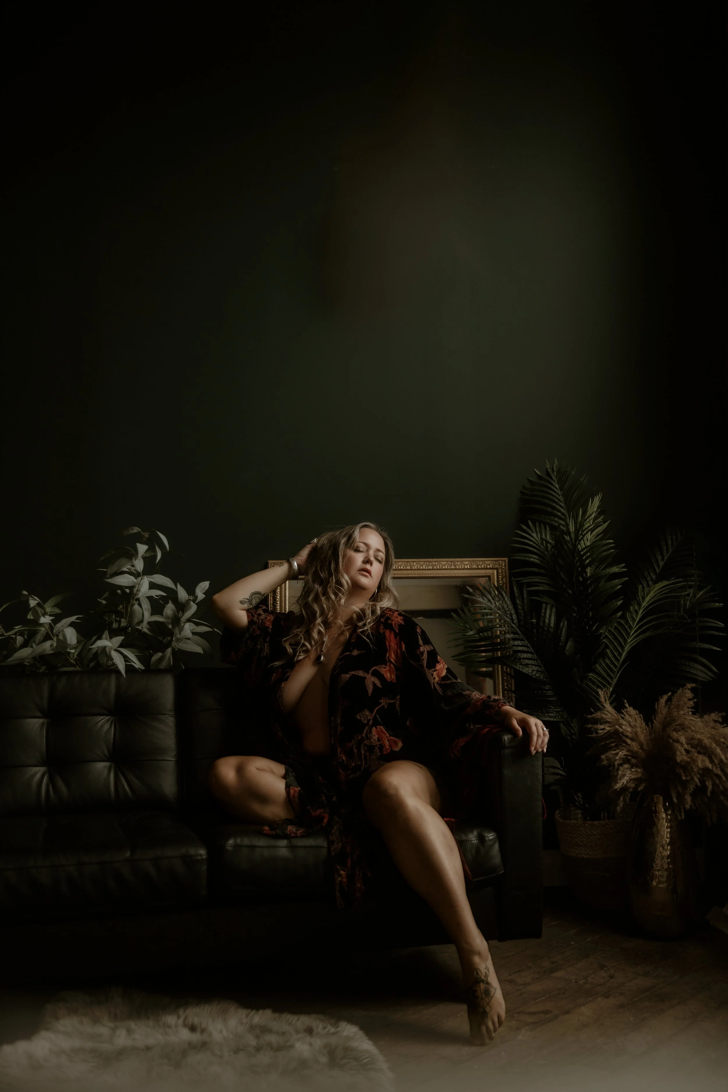A woman with wavy blonde hair seated on a dark leather couch in a dimly lit room, surrounded by indoor plants and a large empty picture frame behind her.