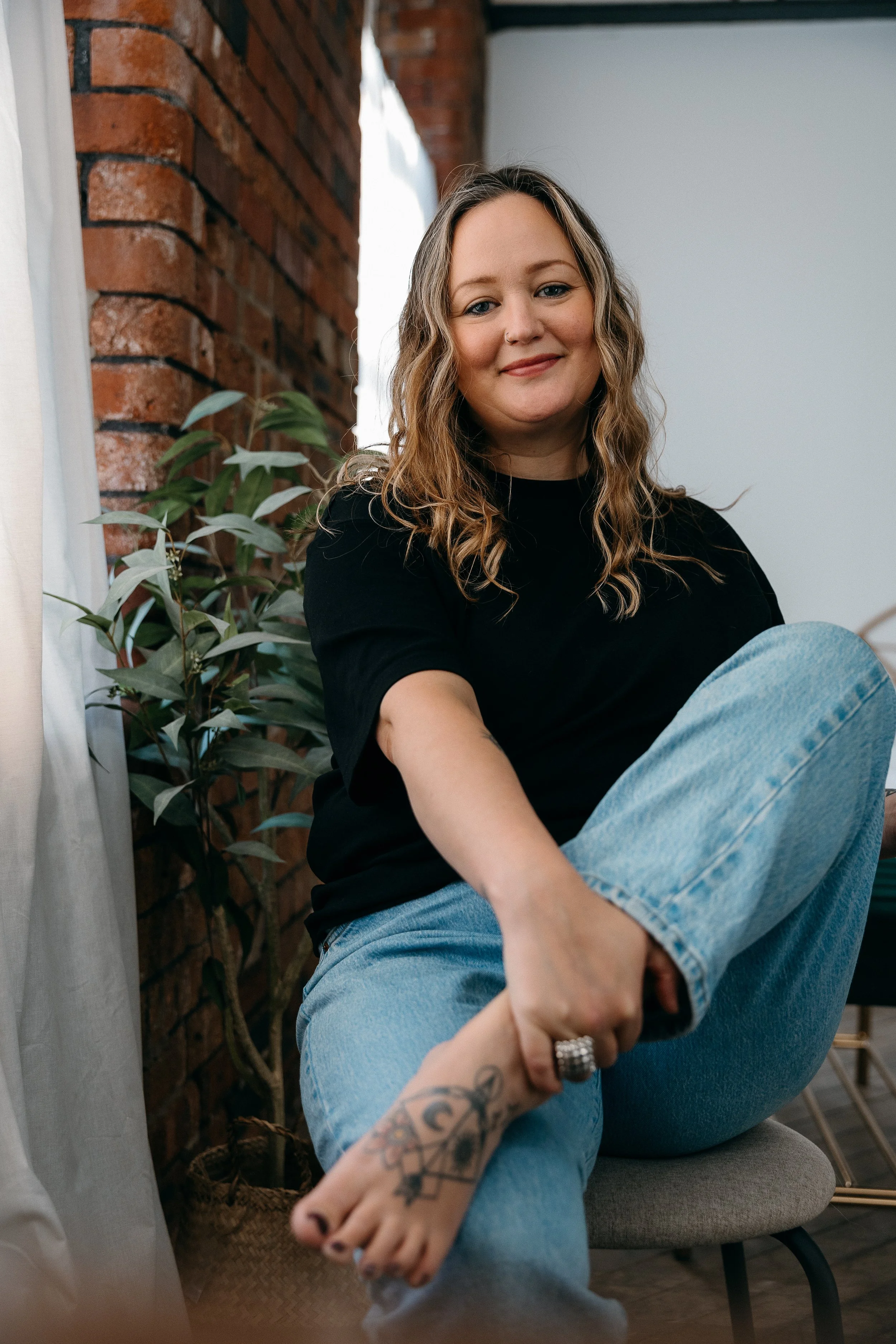 A woman with wavy blonde hair smiling, sitting cross-legged on a chair indoors near a brick wall and a potted plant, wearing a black shirt and light blue jeans.