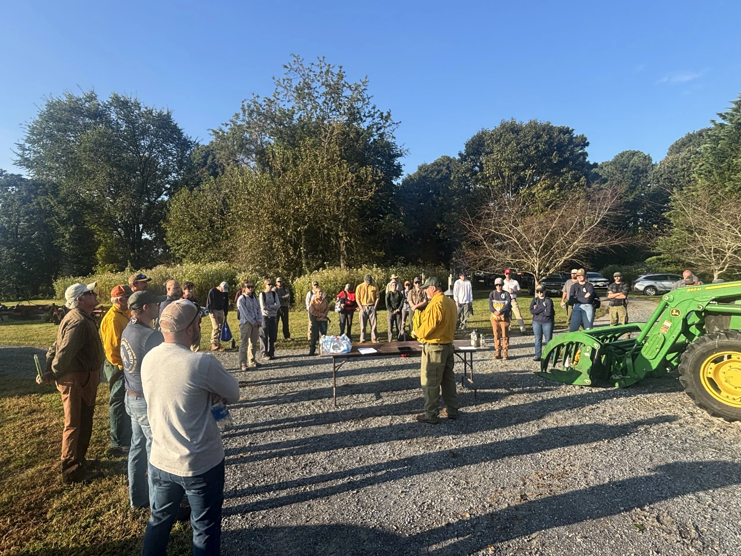 Group of people outdoors listening to a man in a yellow jacket speaking near a table, with a green tractor and parked cars in the background.
