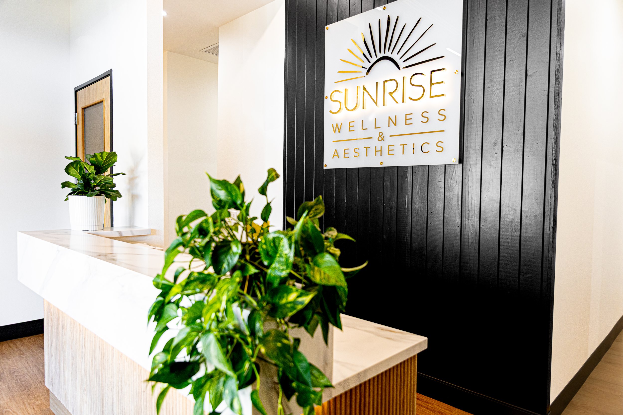 Reception area of Sunrise Wellness & Aesthetics with a white marble counter, green plants, and a sign on a black wooden wall.