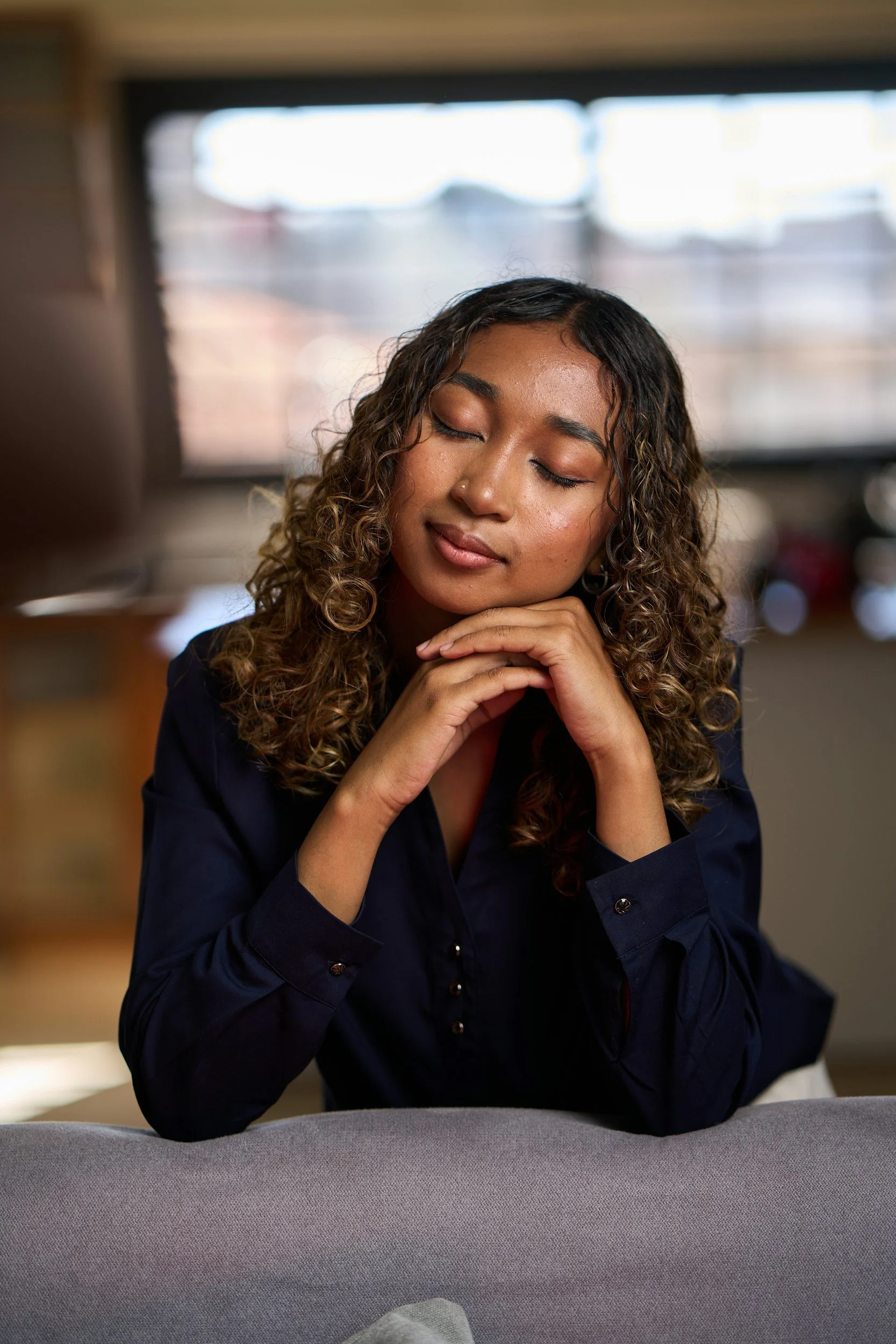 A woman with brown curly hair sitting at a table with her eyes closed and her hands under her chin