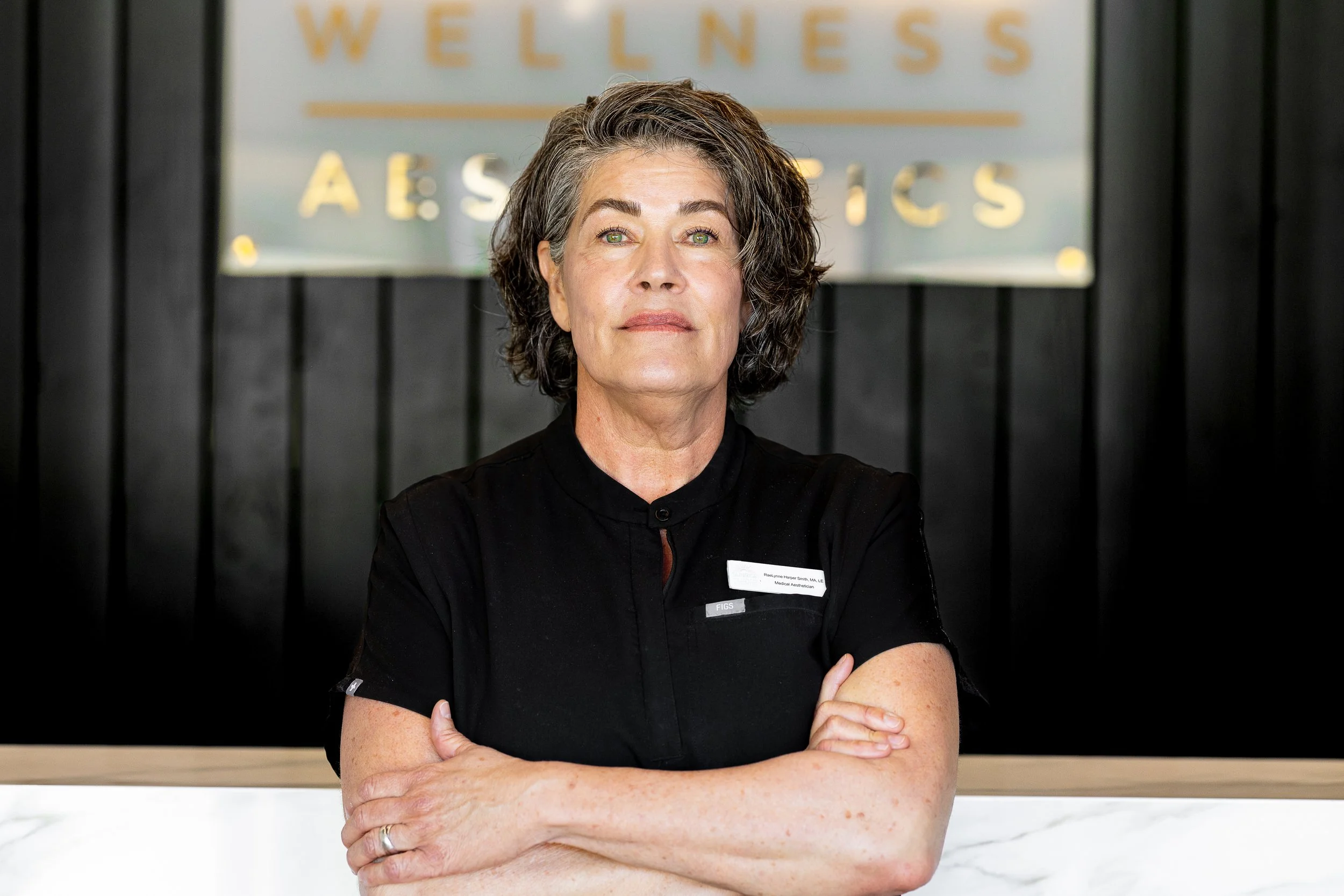 A woman with gray, curly hair, wearing a black medical uniform with her arms crossed, standing in front of a sign that reads 'WELLNESS AESTHETICS' on a black wall background.