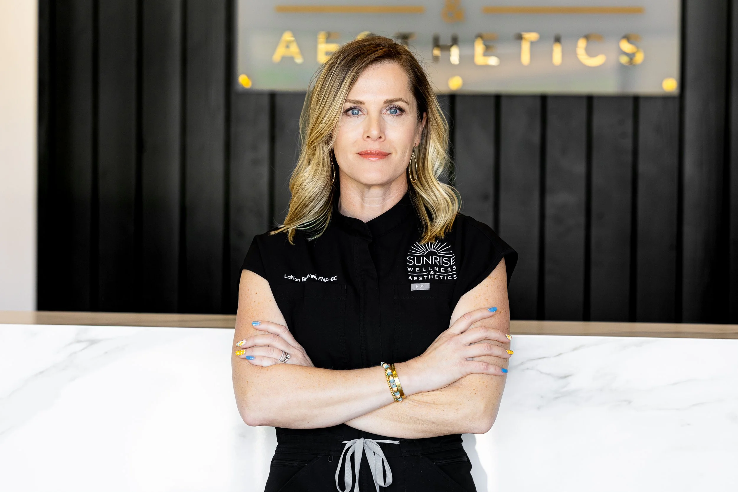 A woman with blonde hair and blue eyes standing with arms crossed, wearing a black vest with 'Sunrise Wellness & Aesthetics' logo in front of a black wooden wall and a white marble counter.