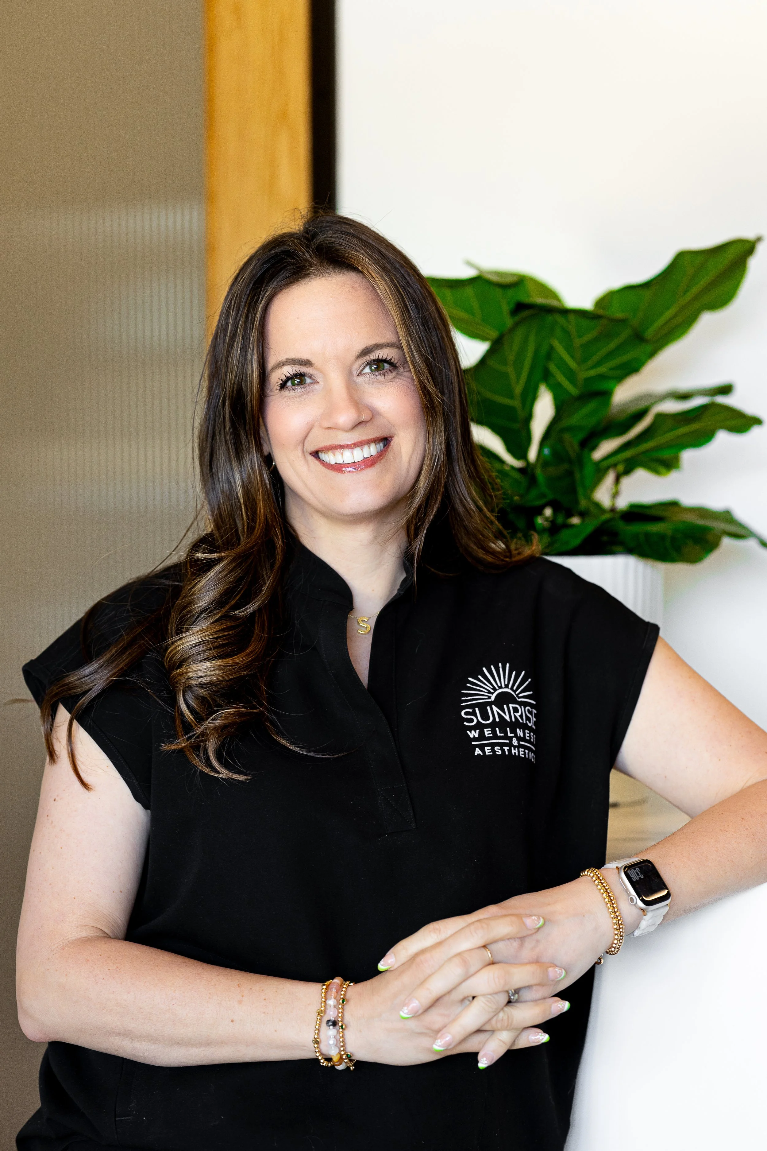 A smiling woman with long, wavy brown hair, wearing a black polo shirt with a logo that reads 'Sunrise Wellness & Aesthetic.' She is standing indoors next to a green plant, with her hands clasped in front of her.