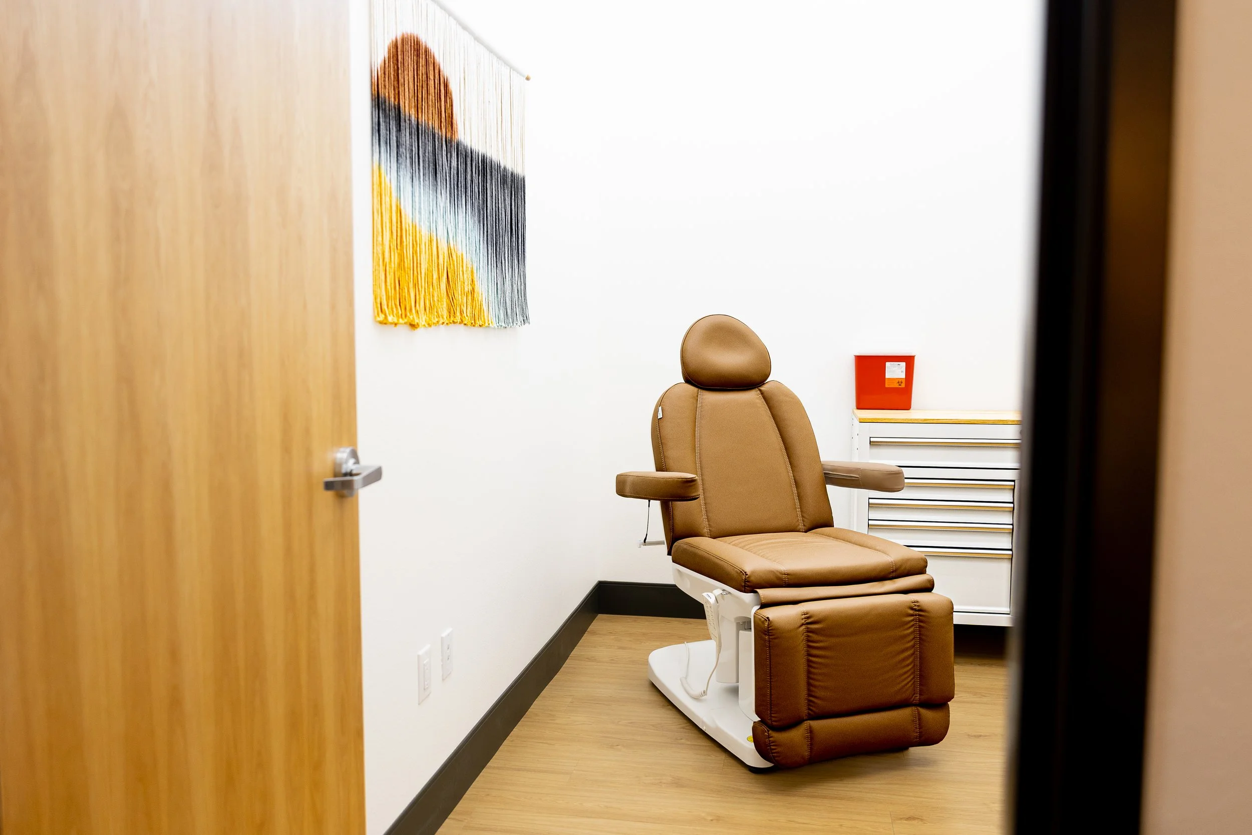 Exam room with a brown medical chair, colorful wall hanging, white wall, and a small white cabinet with a red storage box on top.