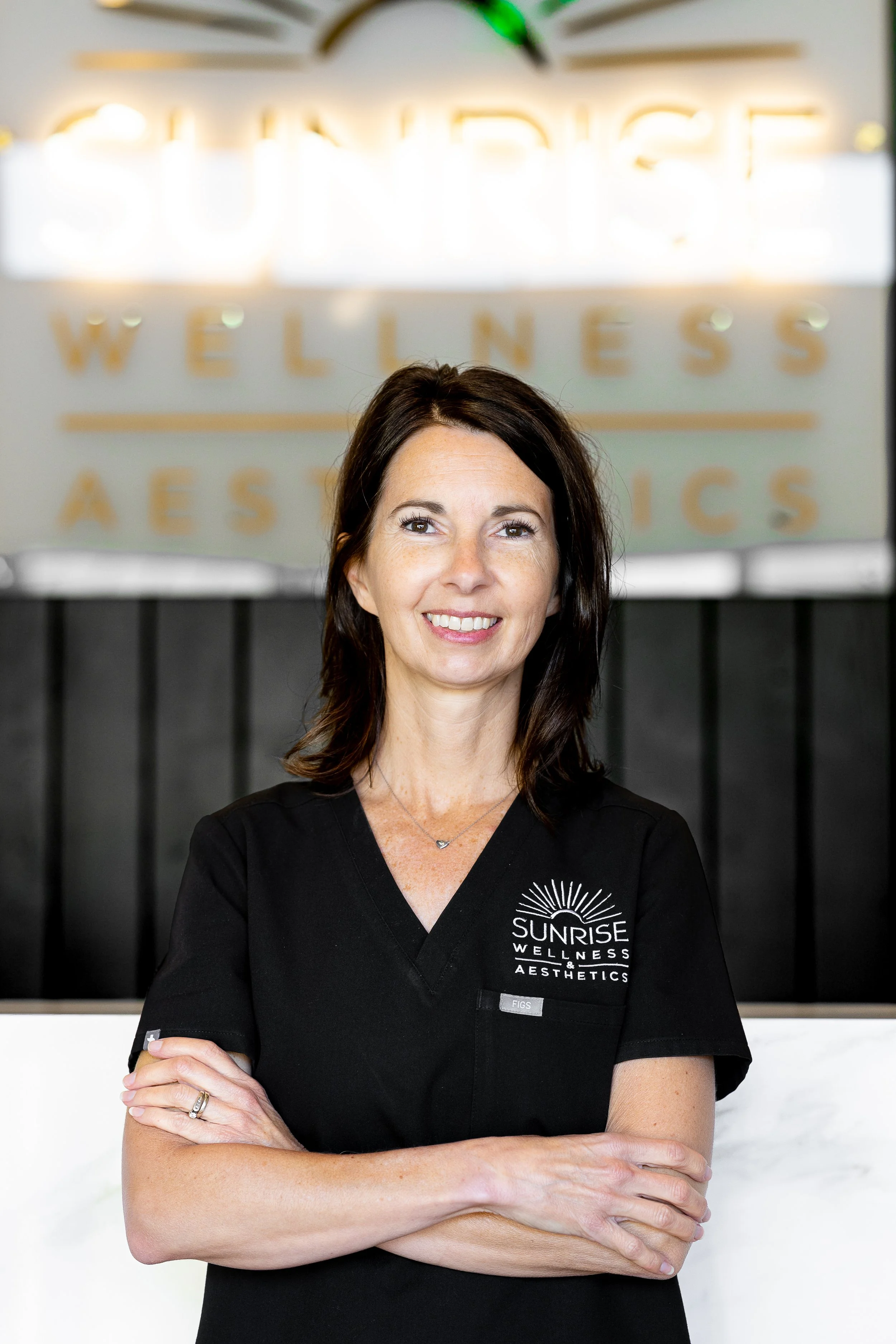 A woman standing with crossed arms, smiling, wearing a black uniform with a 'Sunrise Wellness & Aesthetics' logo, inside a wellness center with a blurred sign in the background.
