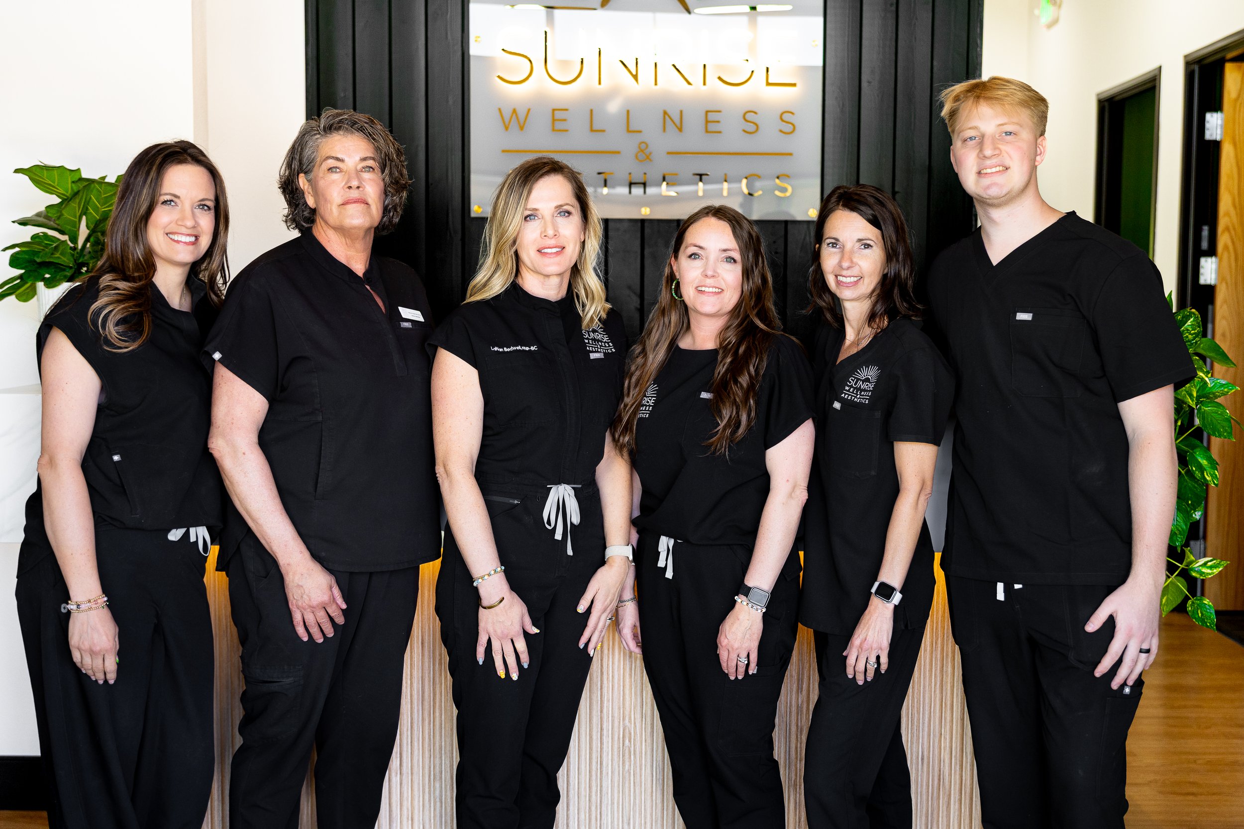 Group of six wellness and aesthetics staff members standing in front of a wellness sign, wearing black scrubs.