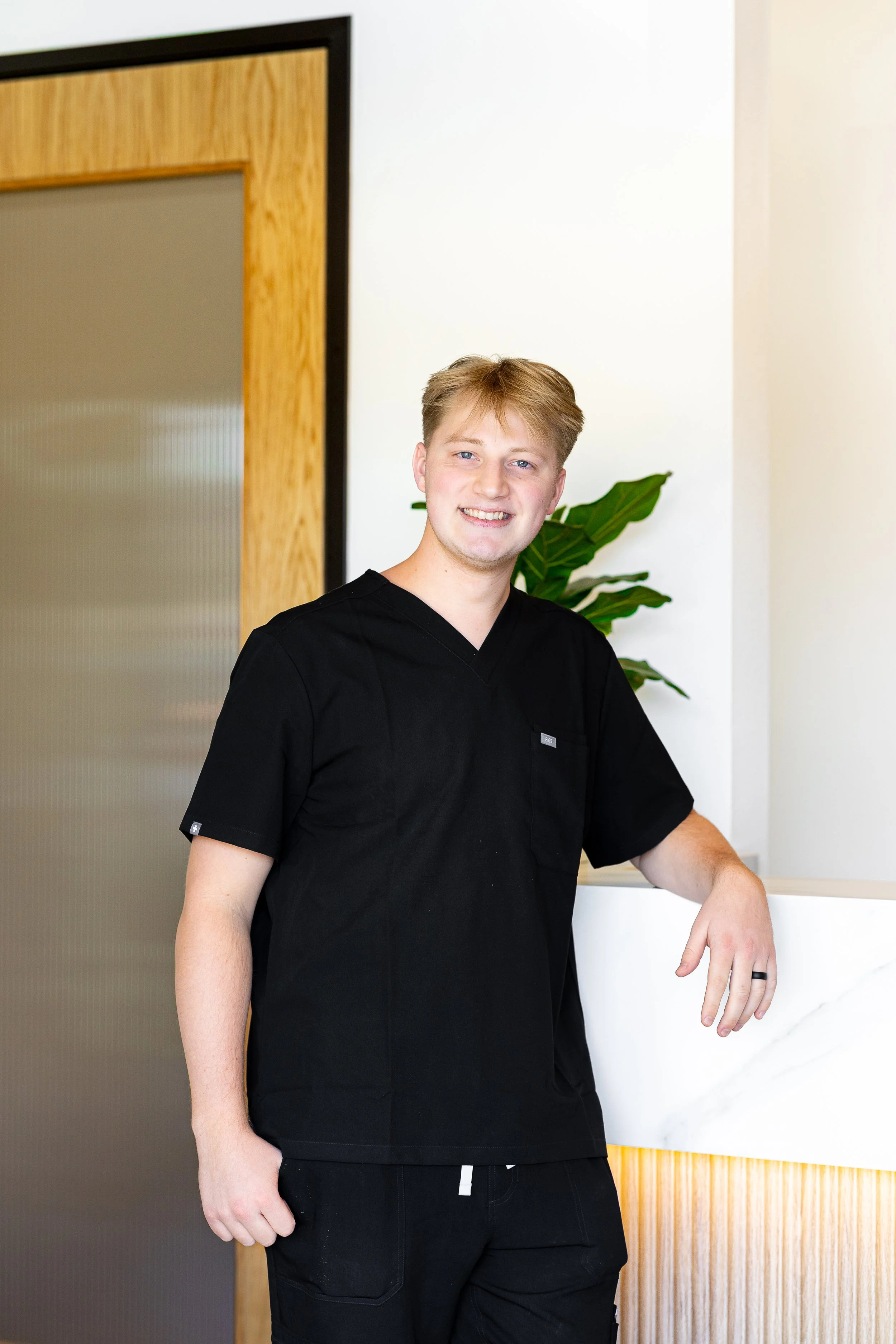 A young man with blonde hair wearing black scrubs, smiling and standing indoors next to a white marble countertop and a green plant, with a wooden door behind him.