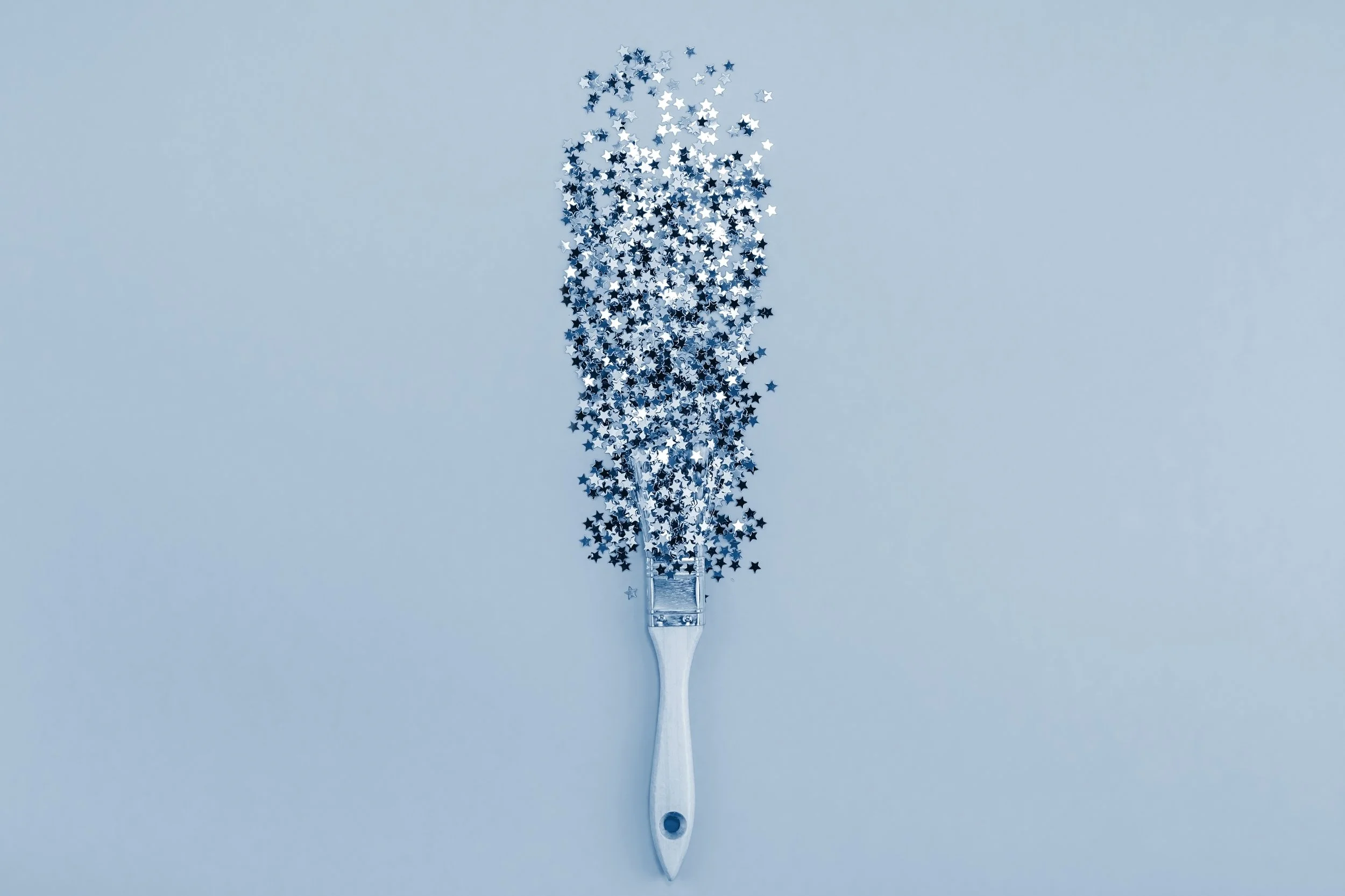A silver utensil with a flat, wide end is covered in silver star-shaped confetti, creating the appearance of a starry spray against a light blue background.