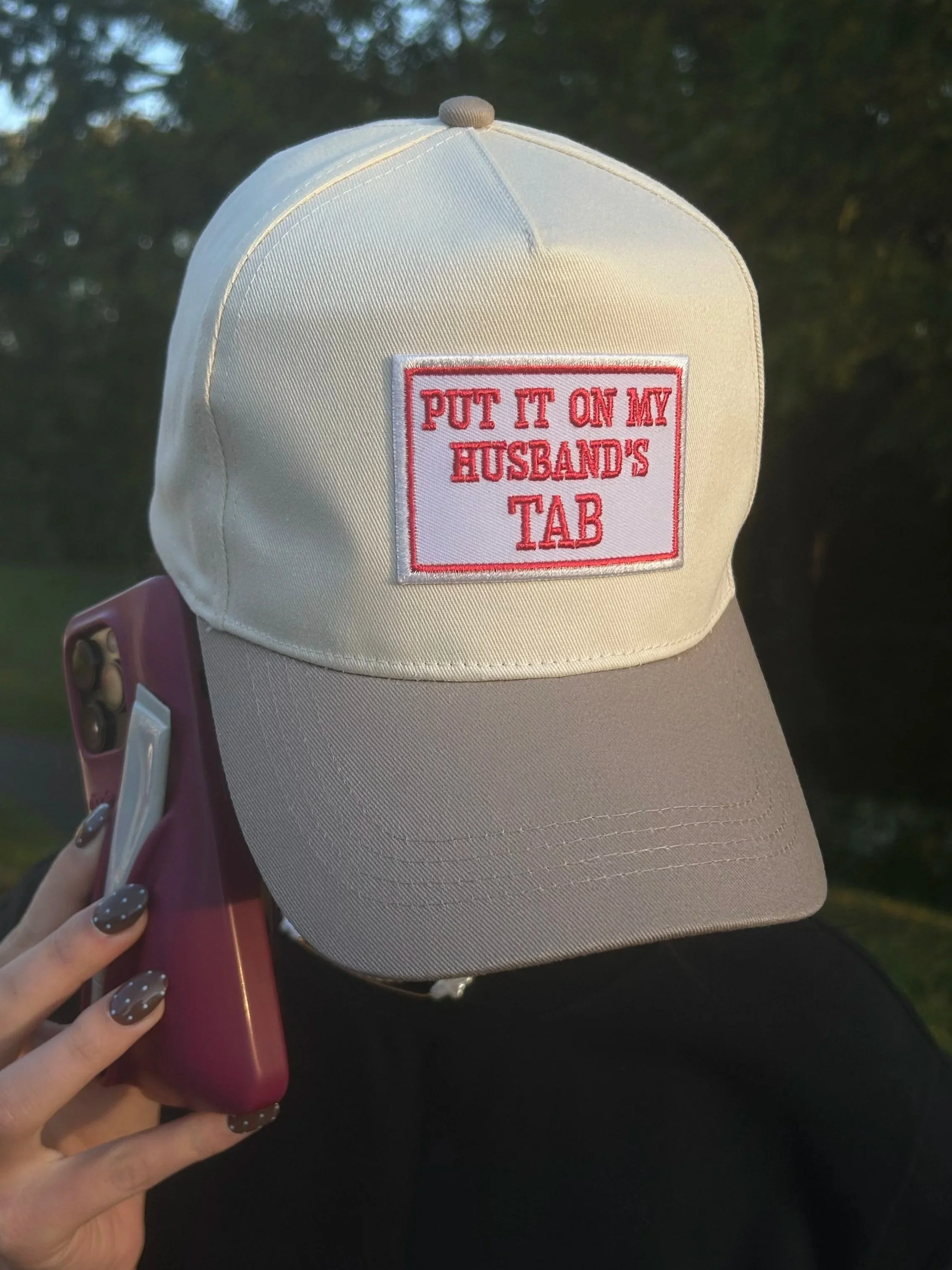 A white and gray baseball cap with a patch that reads, 'PUT IT ON MY HUSBAND'S TAB' in red text, held in a person's hand with fingernails painted polka dot, beside a pink phone, outdoors with trees in the background.