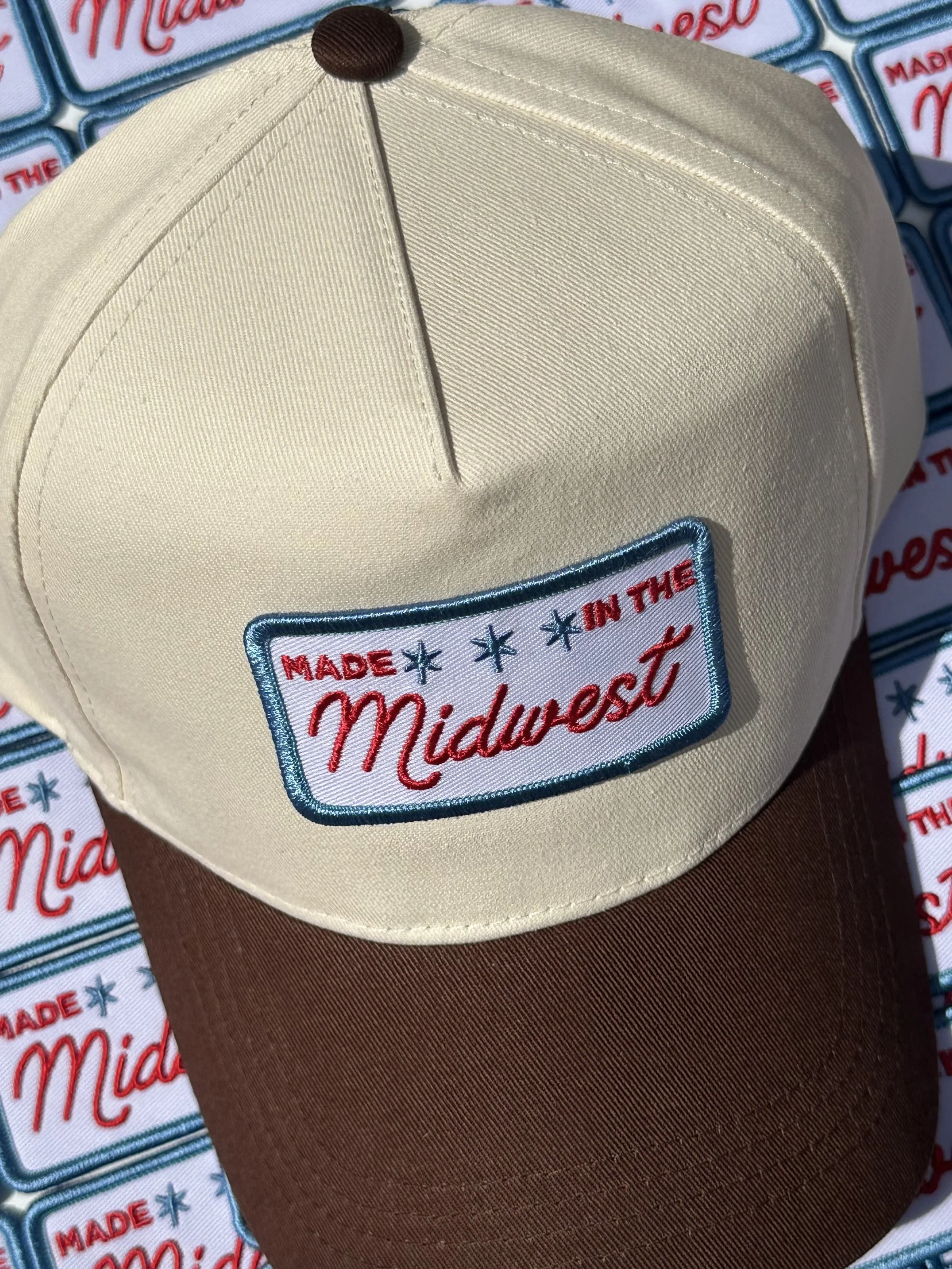 A beige and brown baseball cap with a patch that says 'Made in the Midwest' on the front, placed on a patterned surface with similar patches in the background.