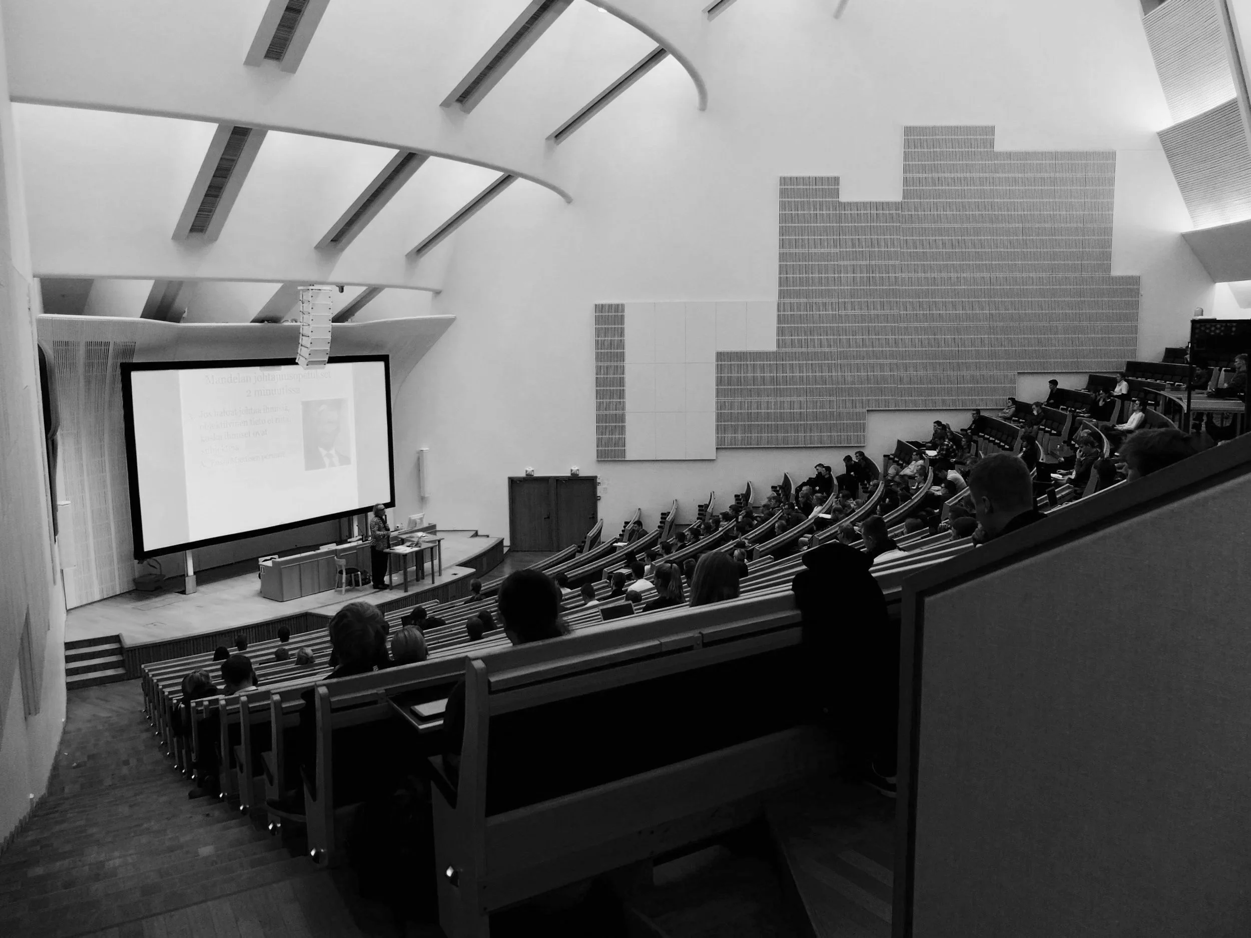 A lecture hall with audience members seated in a curved, tiered arrangement. A speaker stands at a podium in front of a large projection screen displaying a slide with text and a photo. The hall has high ceilings with architectural beams and textured wall panels.