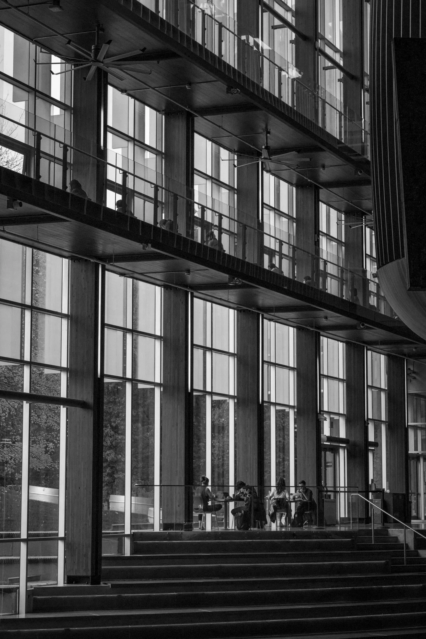 People sitting at tables inside a modern building with large glass windows and multiple floors, captured in black and white.