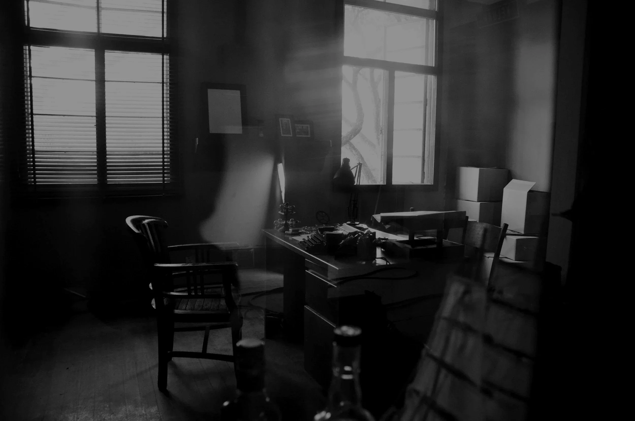 An office or workspace with a desk, chairs, and stacks of boxes, illuminated by sunlight entering through two windows with blinds, creating a moody and shadowy atmosphere.