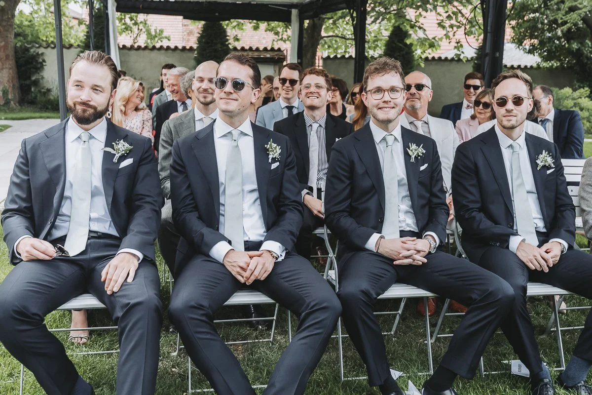 Four men in suits and sunglasses sitting in front row at a wedding or formal event, with other guests seated behind them outdoors.