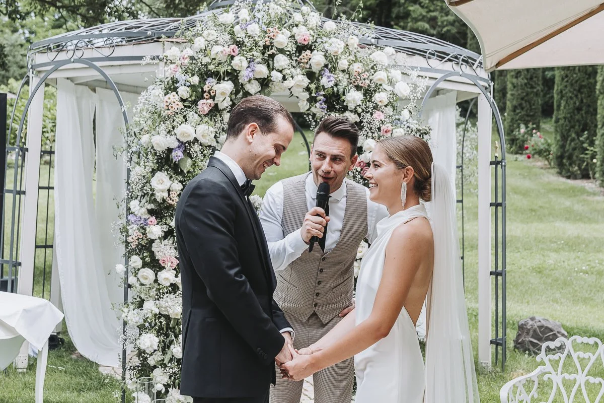 Bride and groom holding hands and smiling during their wedding ceremony outdoors, with an officiant in the middle, under a floral arch with white and pink flowers.