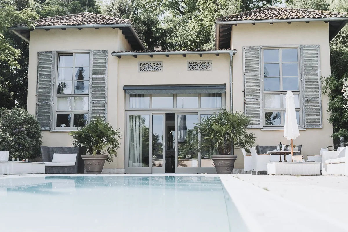 Front view of a house with large windows and shutters, a glass sliding door, a patio with outdoor furniture, and a swimming pool in the foreground.
