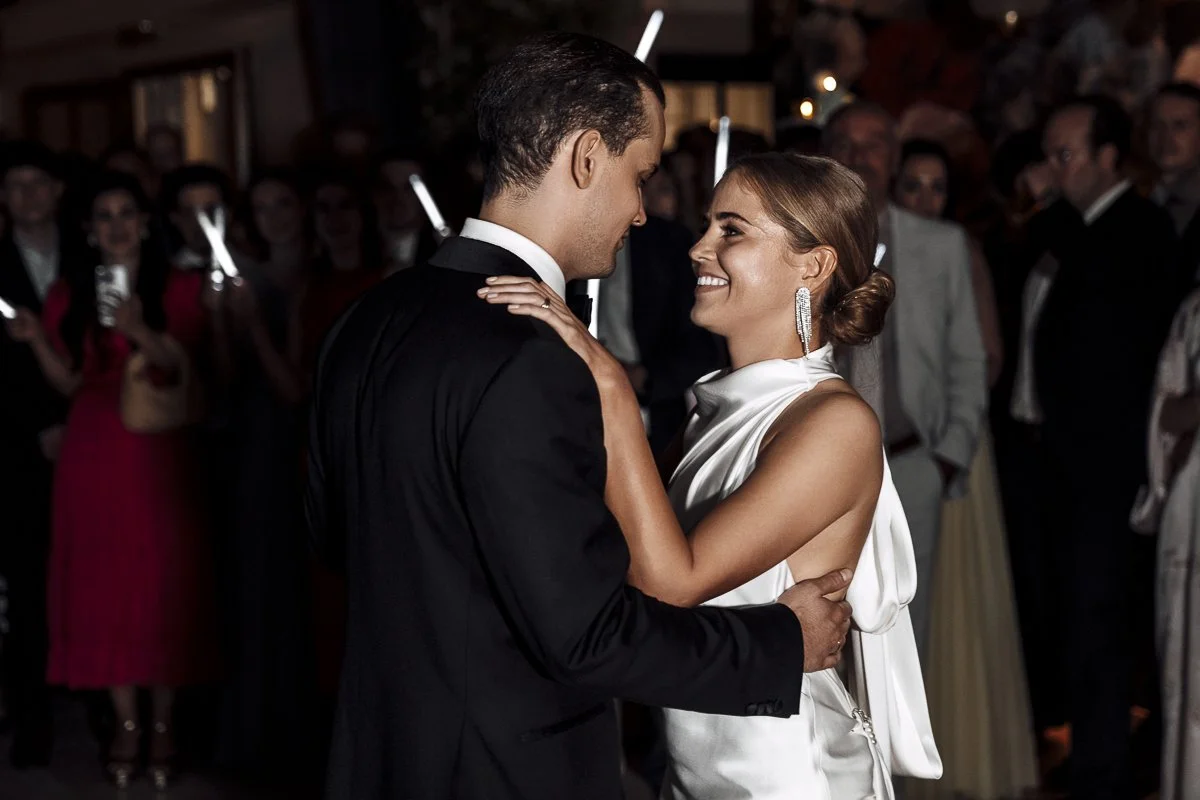 A couple dancing at a formal event, with the woman wearing a white satin dress and the man in a black tuxedo, surrounded by an audience in elegant attire.