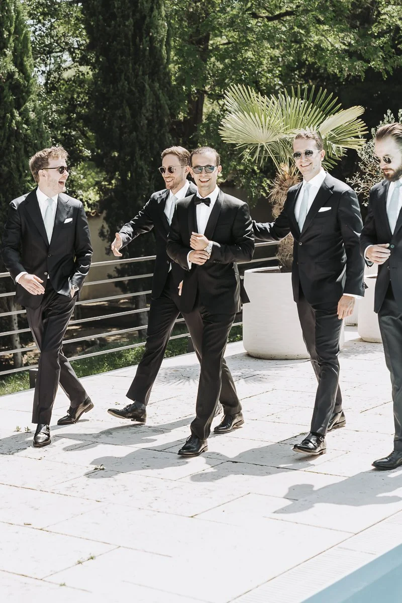 Group of five men in black suits and sunglasses walking outdoors, smiling and chatting. Green trees and potted plants are visible in the background.