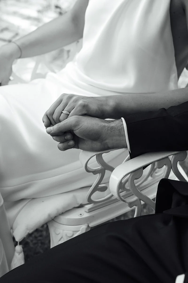 Close-up of a man and woman holding hands, sitting on a decorative white cast iron bench, possibly at a wedding or special event.
