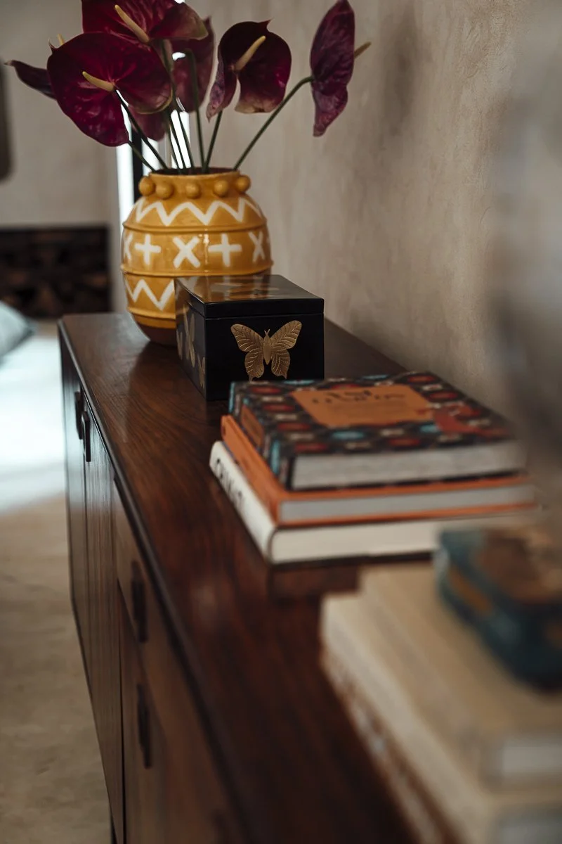 Top of a wooden sideboard or dresser with a vase of purple and pink flowers, a black decorative box with a gold butterfly, and a stack of books.