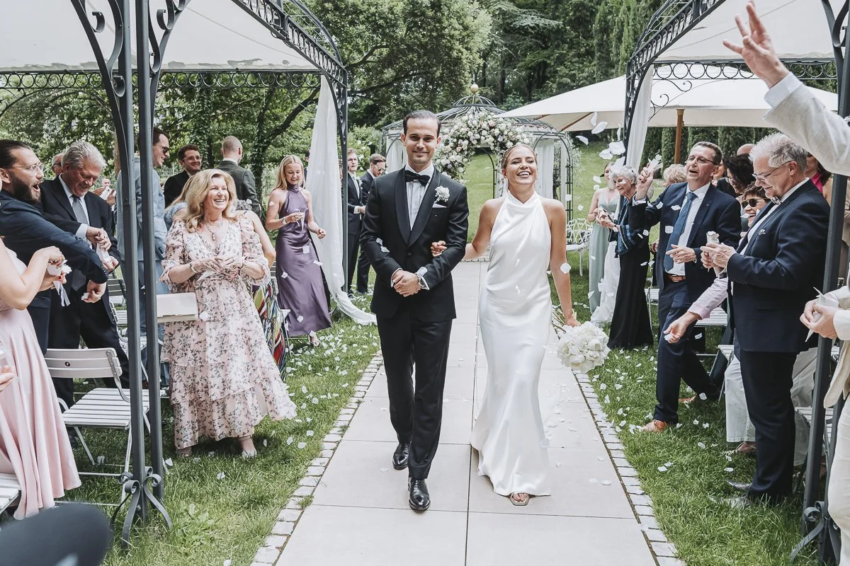 Bride and groom walking down the aisle under a gazebo, smiling, with guests on either side throwing flower petals during an outdoor wedding ceremony.