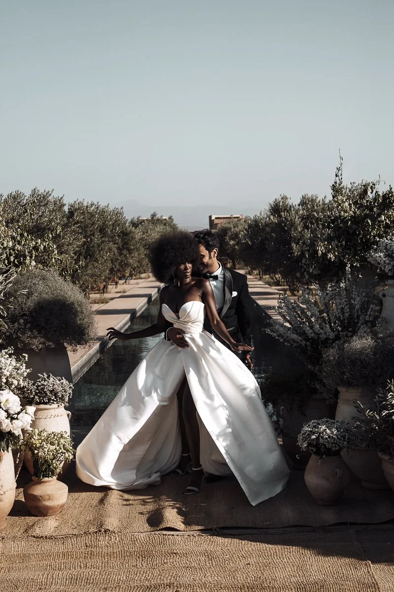 A couple in wedding attire standing outdoors between two garden beds with potted flowers, with a water feature behind them and a clear sky overhead.
