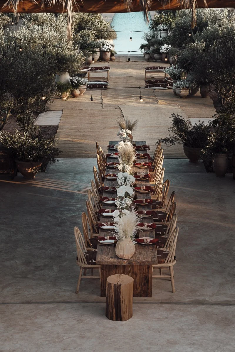 A long rustic dining table set for a gathering, decorated with white floral centerpieces and surrounded by wooden chairs, situated outdoors under string lights with potted plants and trees in the background.