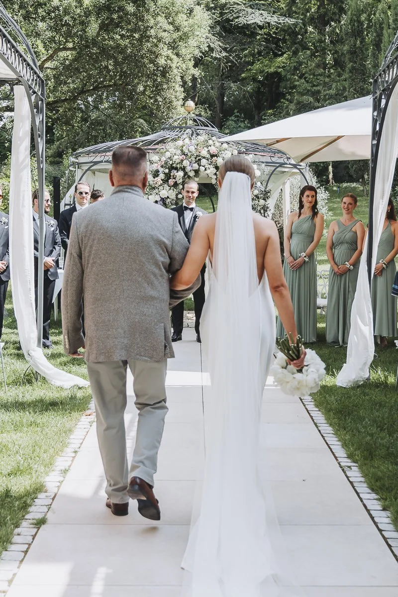 Bride in a white wedding gown with veil holding a bouquet walking down the aisle with her father at an outdoor wedding ceremony, with bridesmaids and groomsmen standing on either side.