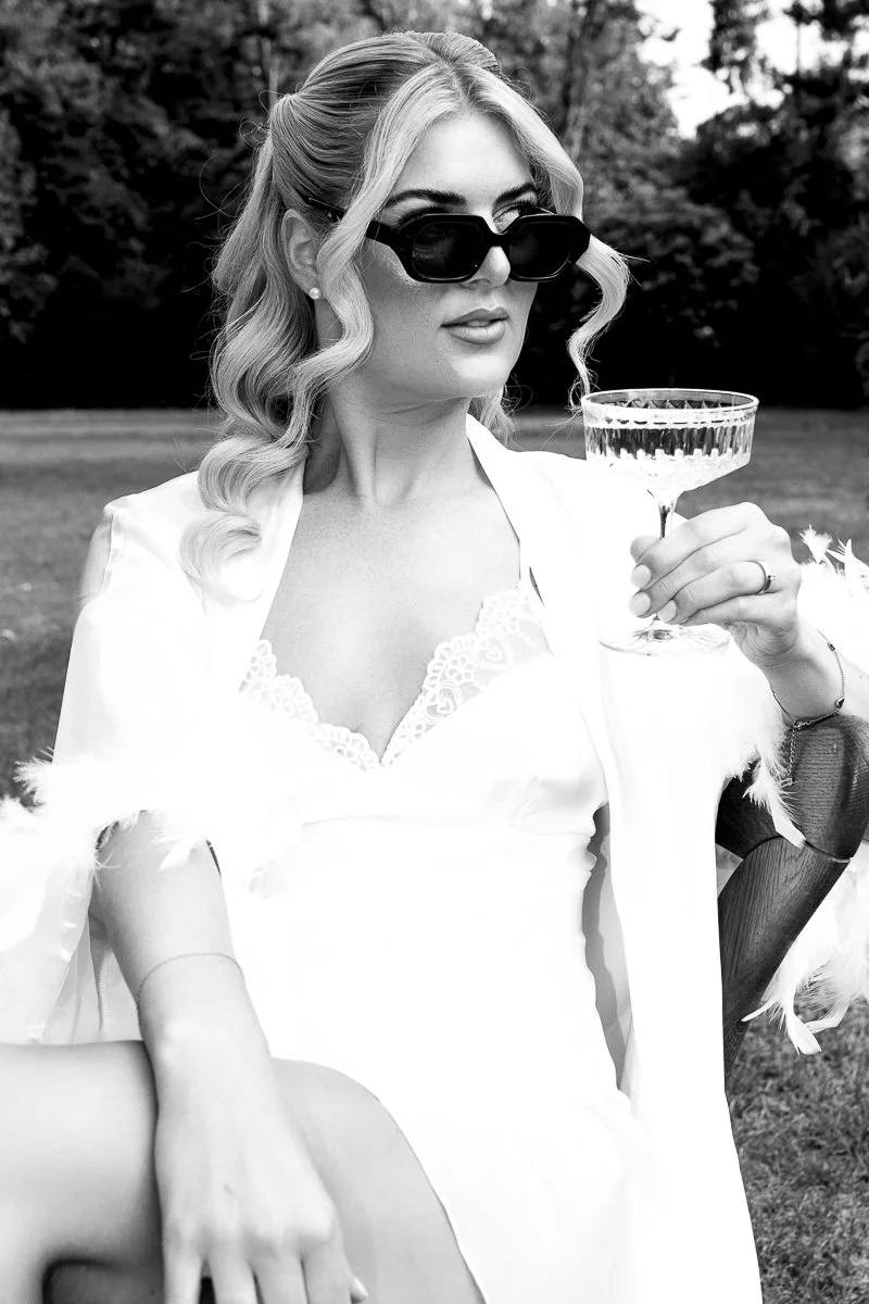 Braut mit Sektglas und Sonnenbrille beim Getting Ready, schwarz-weiß