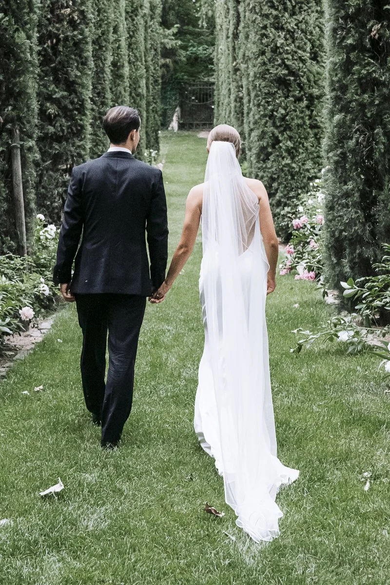 A bride and groom walking hand in hand through a lush, green garden pathway, surrounded by tall trimmed hedges and pink roses.