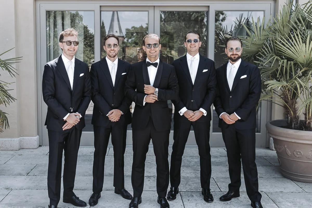 Five men dressed in black tuxedos, white shirts, and sunglasses, standing outdoors in front of glass doors, with potted plants on each side, smiling.