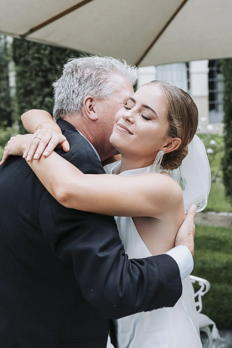 A bride and an older man, likely her father, hugging each other at a wedding outdoors underneath a large umbrella.