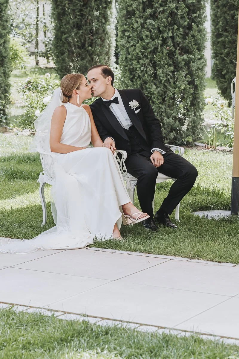 A bride and groom sitting on a white bench, sharing a kiss outdoors surrounded by greenery.