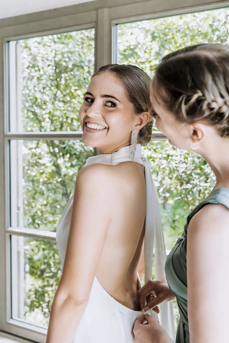 A smiling woman in a white dress is having her dress fastened by another woman who is wearing a green dress. They are inside a room with large windows showing green trees outside.