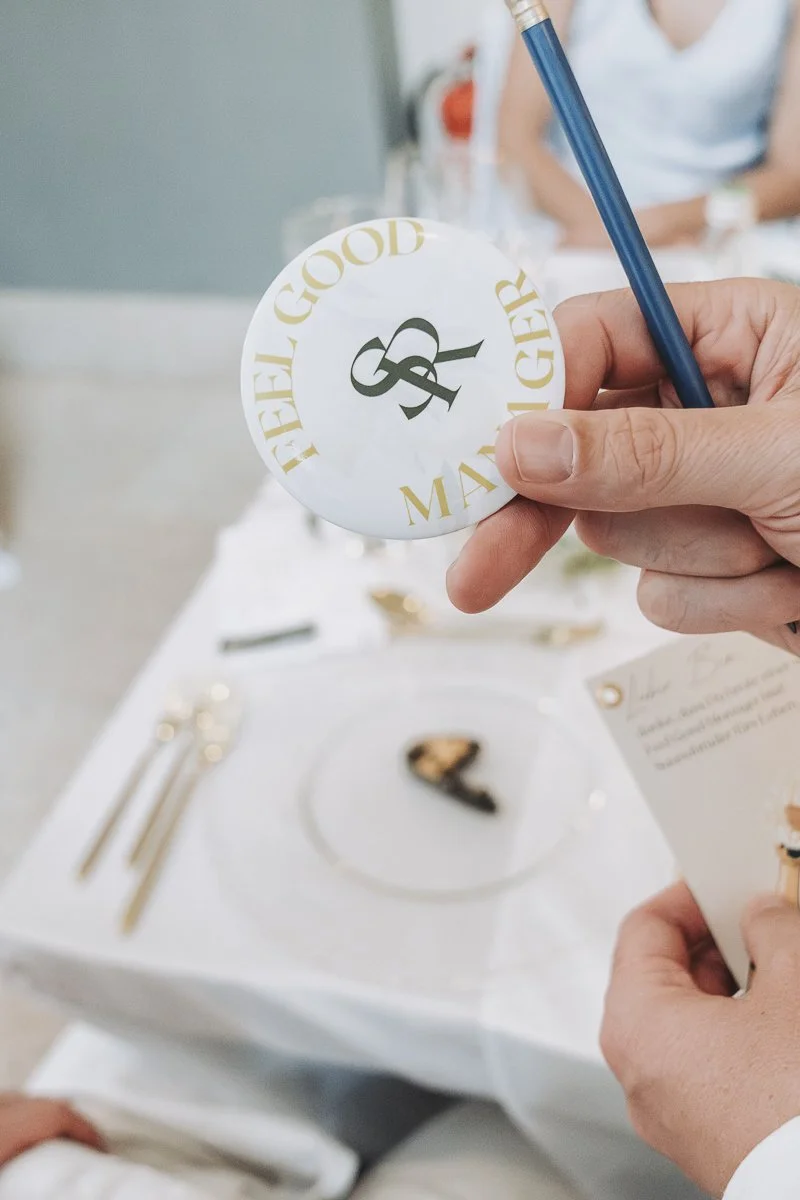 Person holding a paper coaster with the words "Feel Good Greater" and a stylized ampersand, over a dining table set with silverware and a small piece of cake.