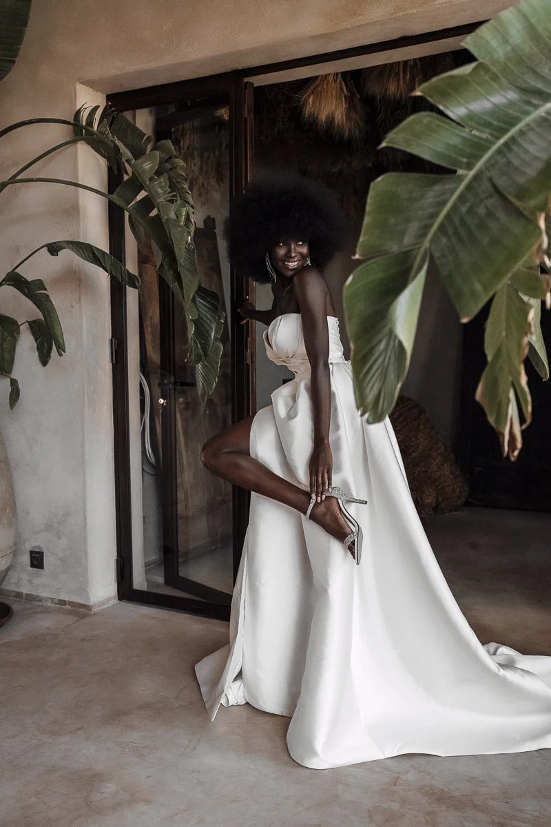 A woman in a strapless white wedding gown lifting her leg to show her high-heeled shoe, smiling in a lush, indoor setting with large green leaves.