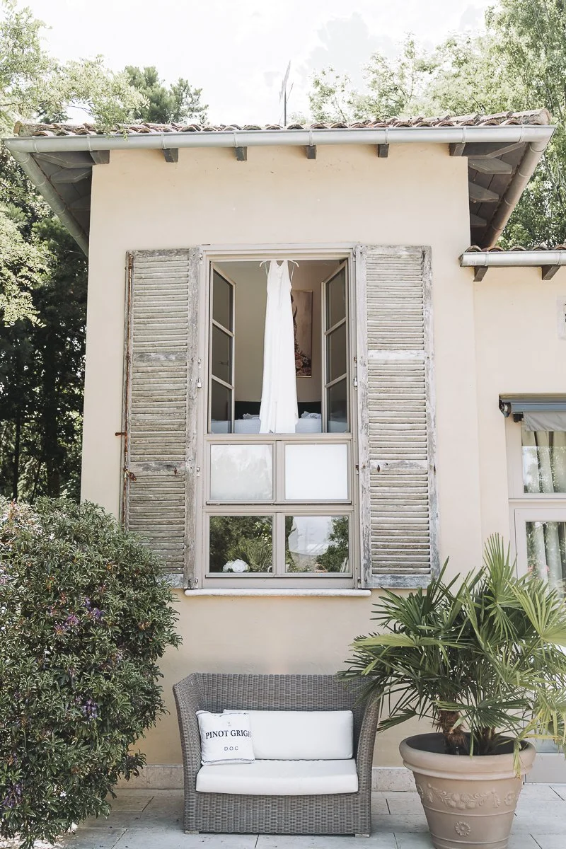 The exterior wall of a house with an open window, displaying a hanging white curtain inside. There is a wicker bench with a white cushion and a pillow that reads 'PINOT GRIGIO' in front of the house, with greenery and a potted plant nearby.