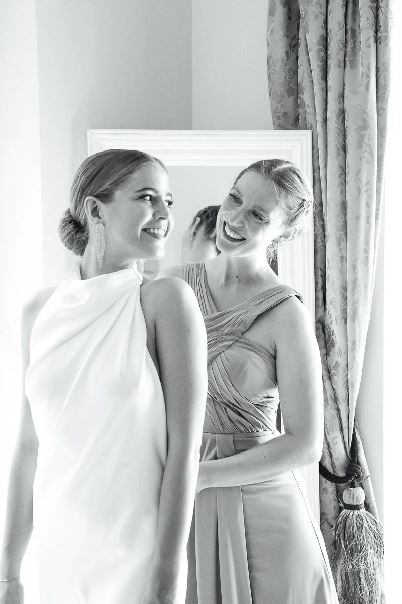Two women smiling and looking at each other, one in a white dress and the other in a light-colored dress, standing near a mirror with patterned curtains in the background.