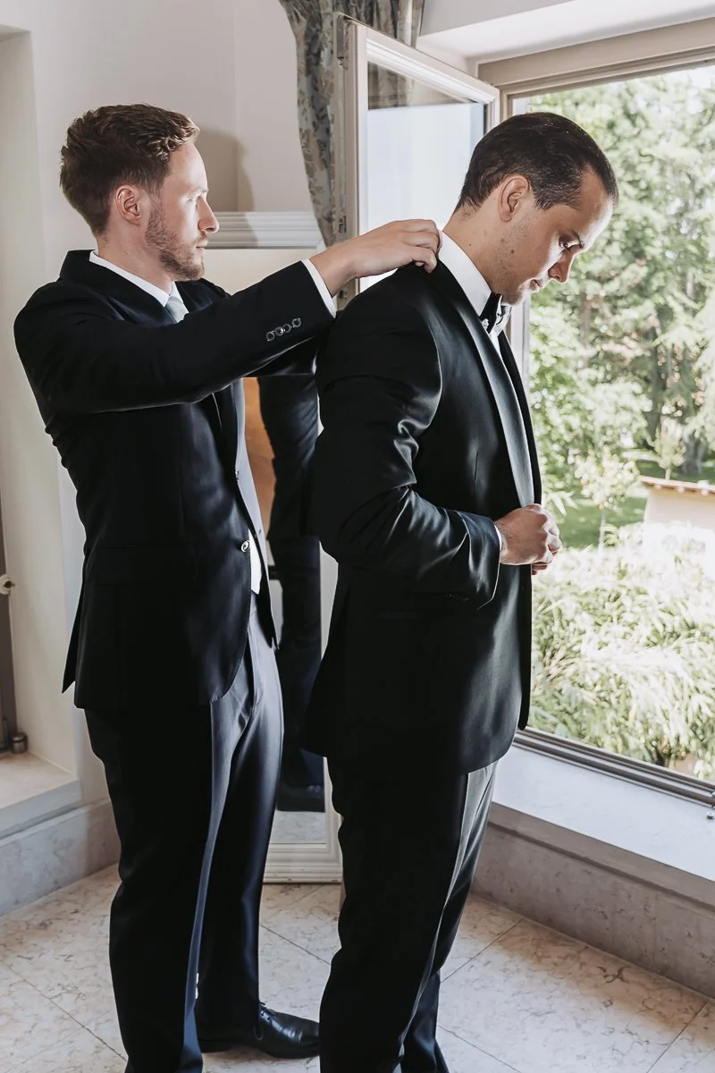 Two men in tuxedos getting ready, one helping the other with his jacket near a window with outdoor greenery visible.