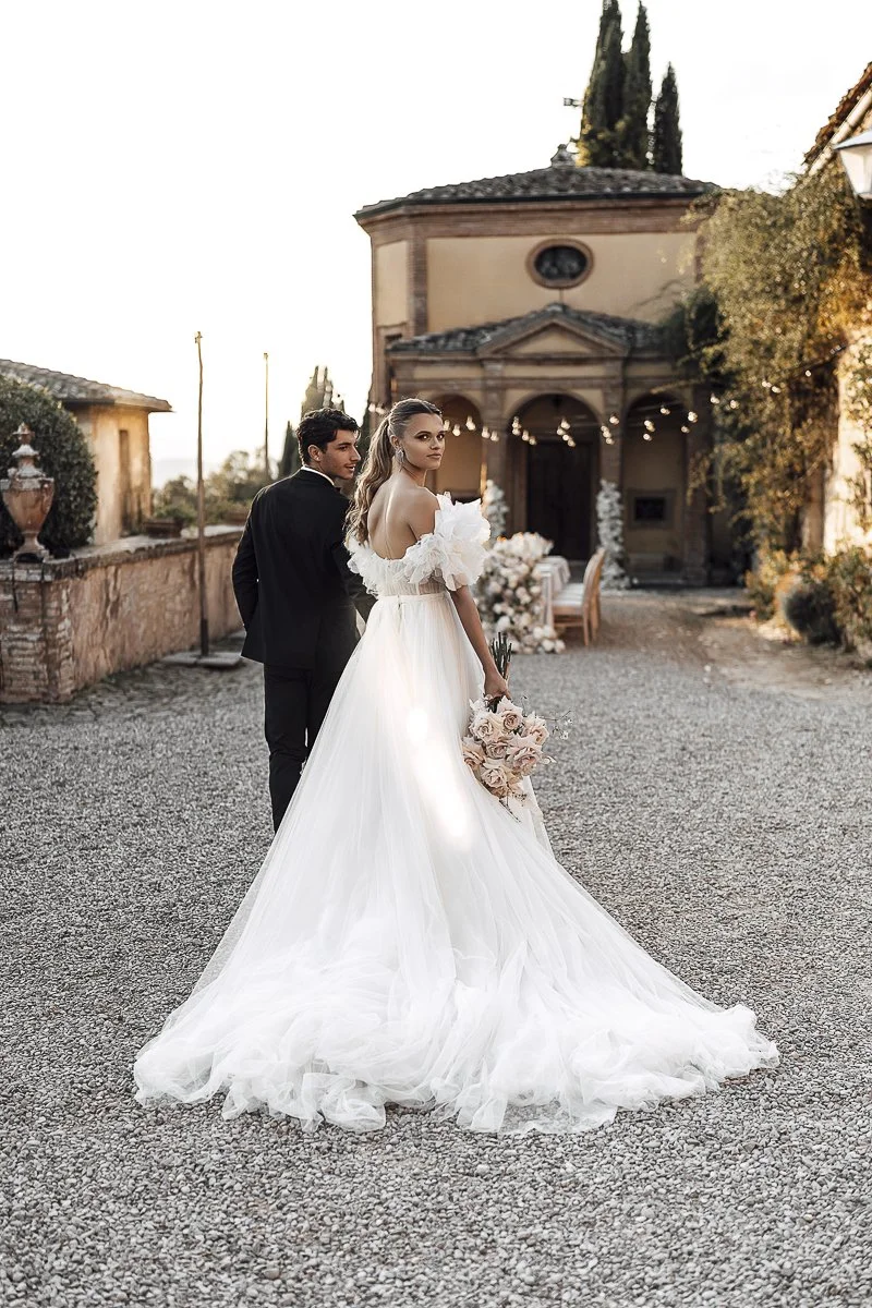 A bride in a white wedding gown holding a bouquet of pink roses, standing next to a groom in a black tuxedo, on a gravel pathway outside a building with string lights at sunset.