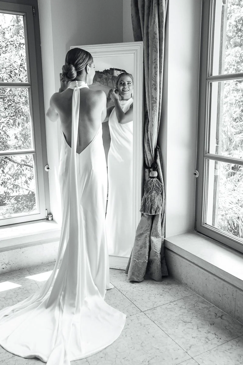 A woman in a backless white gown looking at herself in a full-length mirror in a bright room with windows and curtains.