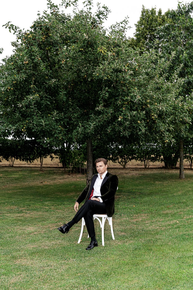 Bräutigam im Smoking auf weißem Stuhl unter Obstbäumen vor der Gartenhochzeit