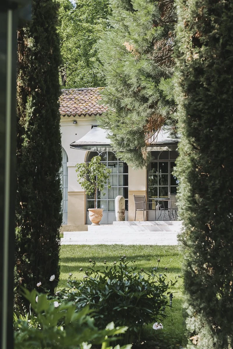 View of a house backyard seen through tall, dense greenery including trees and bushes, with a patio that has chairs, a table, and decorative items.