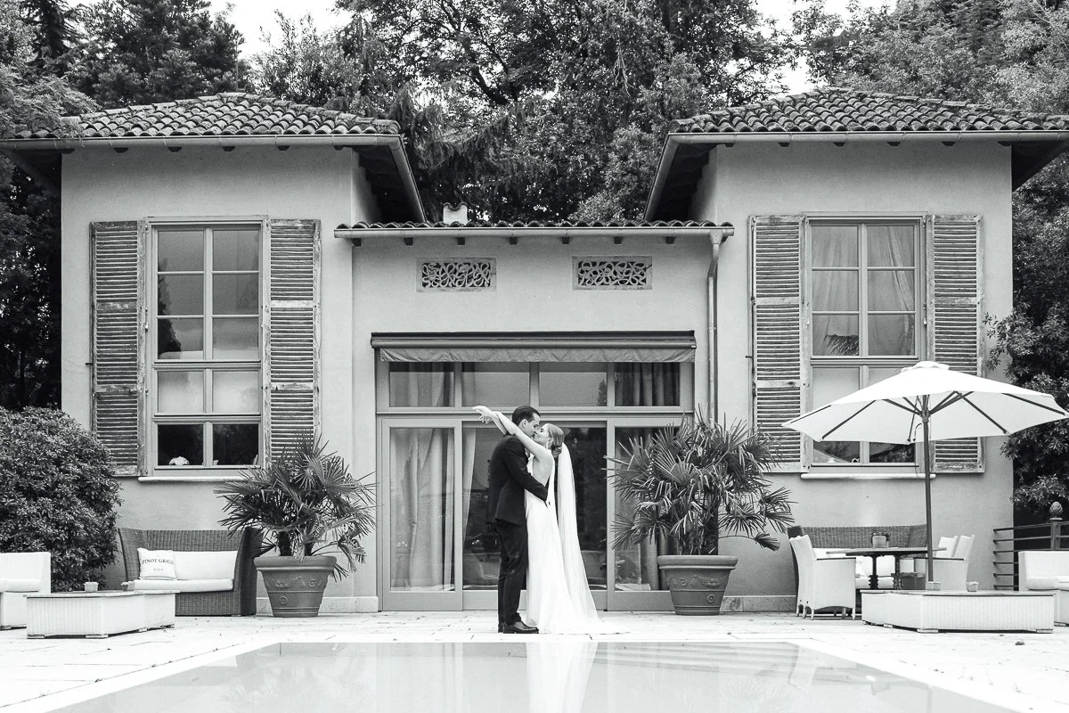 Couple in wedding attire sharing a kiss in front of a house with large windows, shutters, and potted plants, near a swimming pool with outdoor furniture and umbrella.