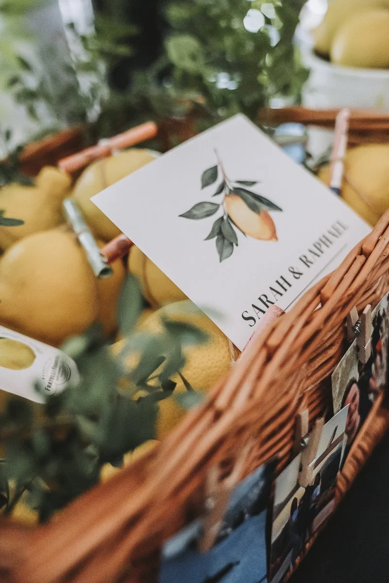 A close-up of a basket filled with yellow apples and a card with an illustration of a peach and leaves, with the names Sarah and Raphael written on it.