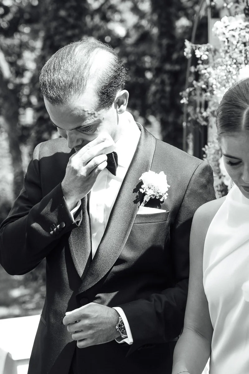 A man in a tuxedo with a boutonniere, wiping his eyes during a wedding ceremony.