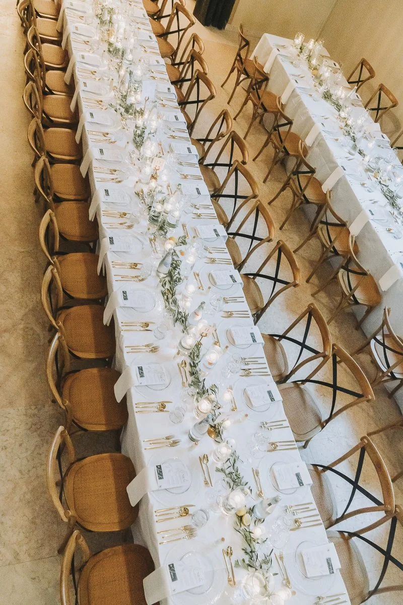 A long banquet table set for a formal event, with white tablecloths, gold cutlery, glassware, candles, and greenery decorations.